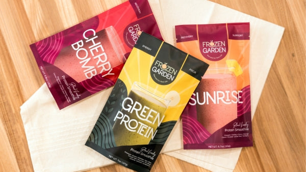 Three pre-packaged smoothie pouches from the brand "Frozen Garden" are arranged on a cream-colored cloth over a light wooden surface.