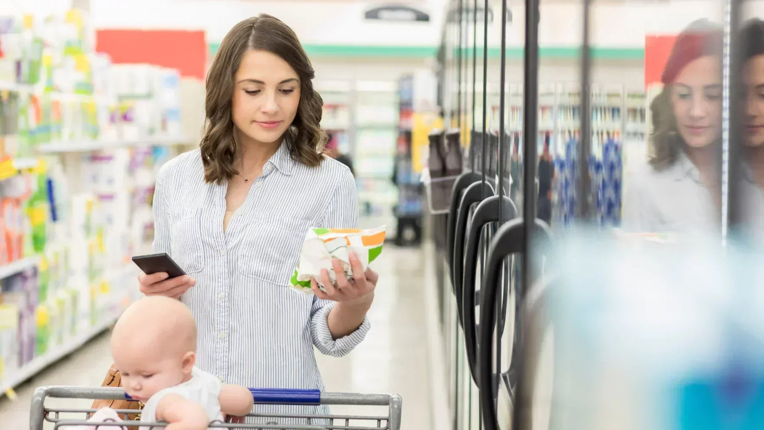 a woman look at a nutrition label with a baby in shopping cart