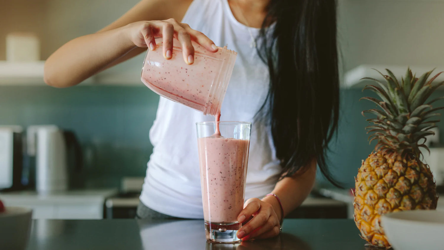 A woman pouring a pink smoothie from a blender pitcher into a glass, with a pineapple in the background, depicting the convenience of maintaining healthy eating habits through nutrient-rich smoothies.