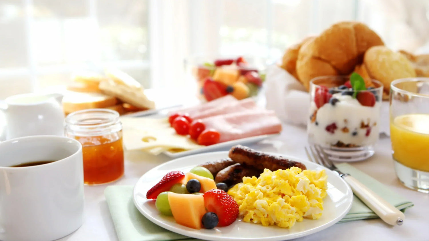A full breakfast table setting featuring eggs, sausage, fresh fruit, coffee, juice, and pastries.