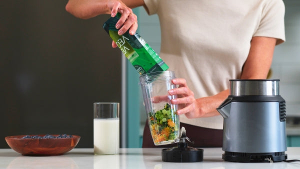 A person pouring a ready-to-blend green smoothie  into a blender cup on a kitchen counter, with a glass of milk and a bowl of blueberries nearby.