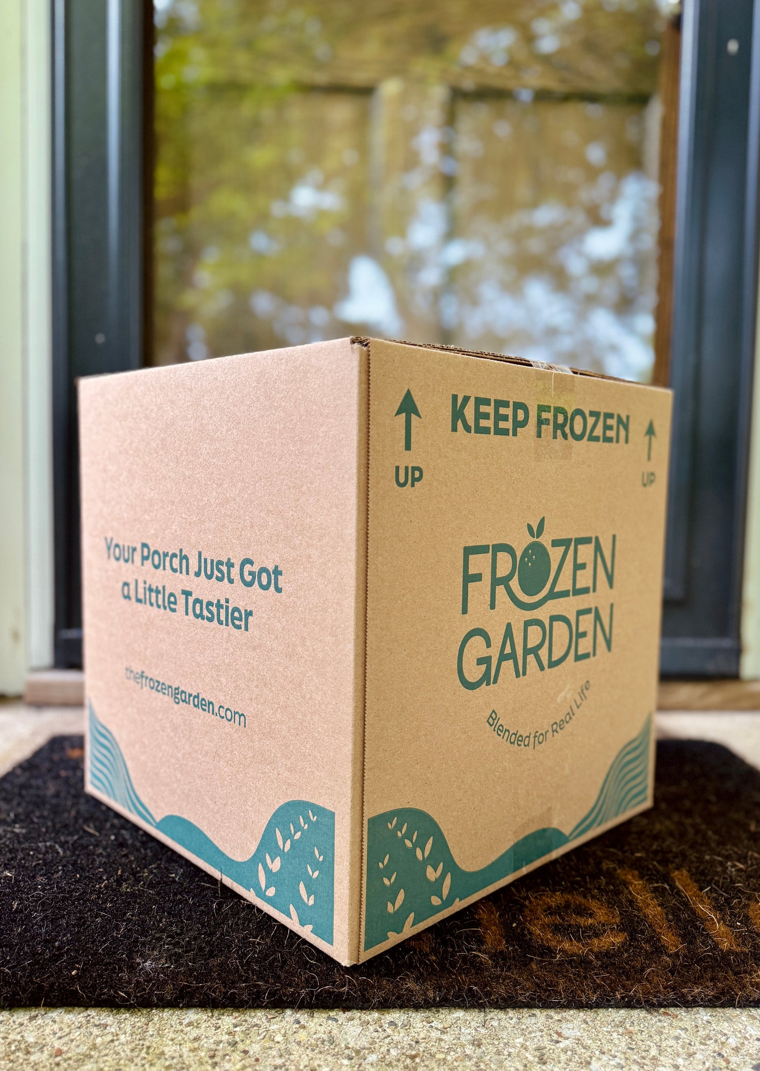 A medium-sized, square cardboard delivery box sits on a dark "hello" doormat in front of a glass front door. The box features dark teal printed text and graphics for Frozen Garden.