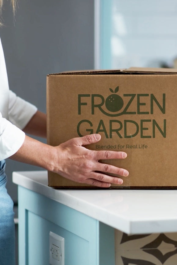 Person holding a cardboard box labeled 'Frozen Garden' in a kitchen.