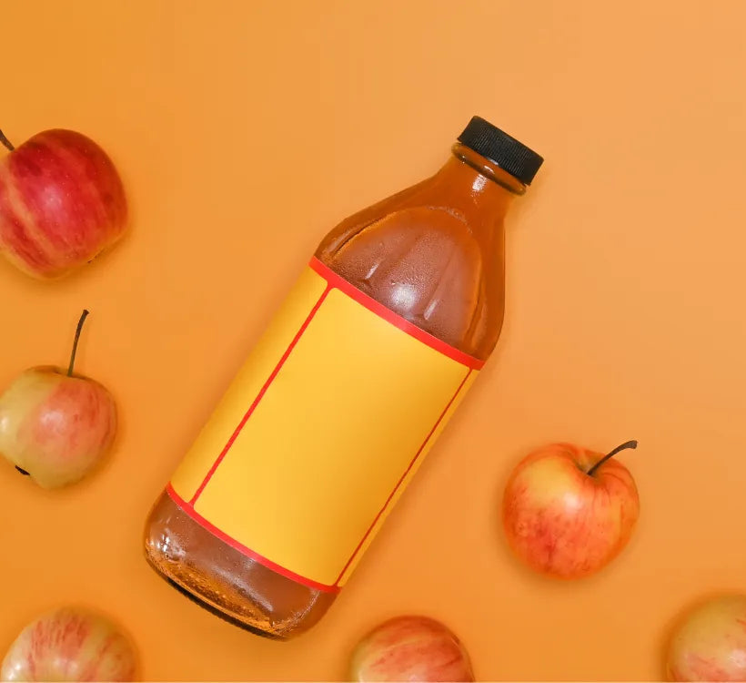 A top-down view of a glass bottle filled with apple cider vinegar, featuring a blank yellow label and surrounded by scattered apples on an orange background