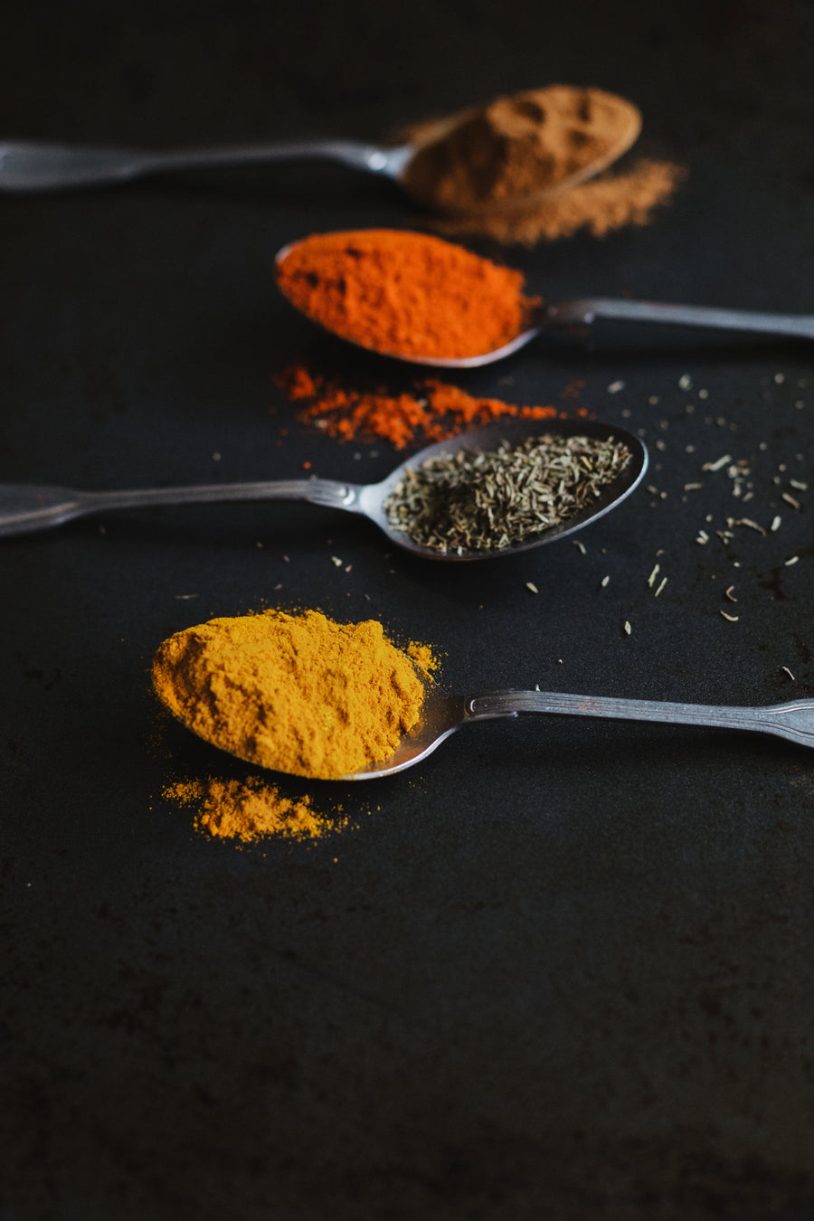 Four metal spoons arranged on a dark surface, each holding a different spice