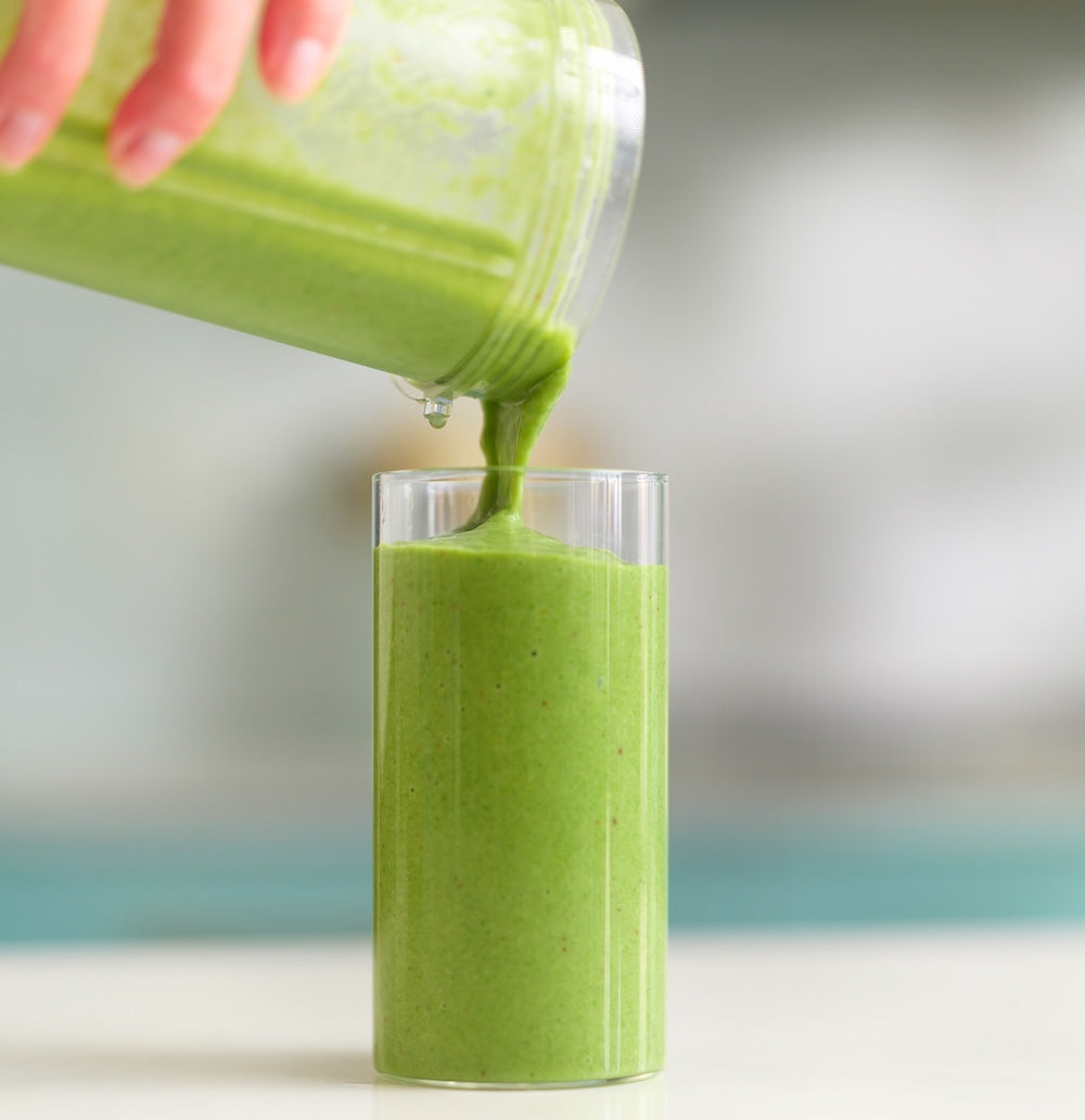 Green smoothie being poured from a blender into a glass on a white surface.