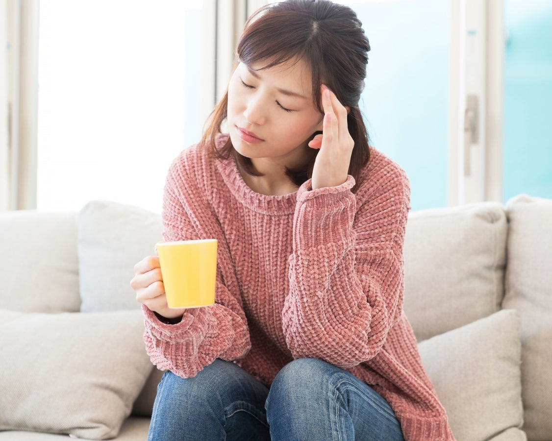 Woman sitting on a couch holding a yellow mug and touching her temple in pain, illustrating common dehydration symptoms like headaches and fatigue.