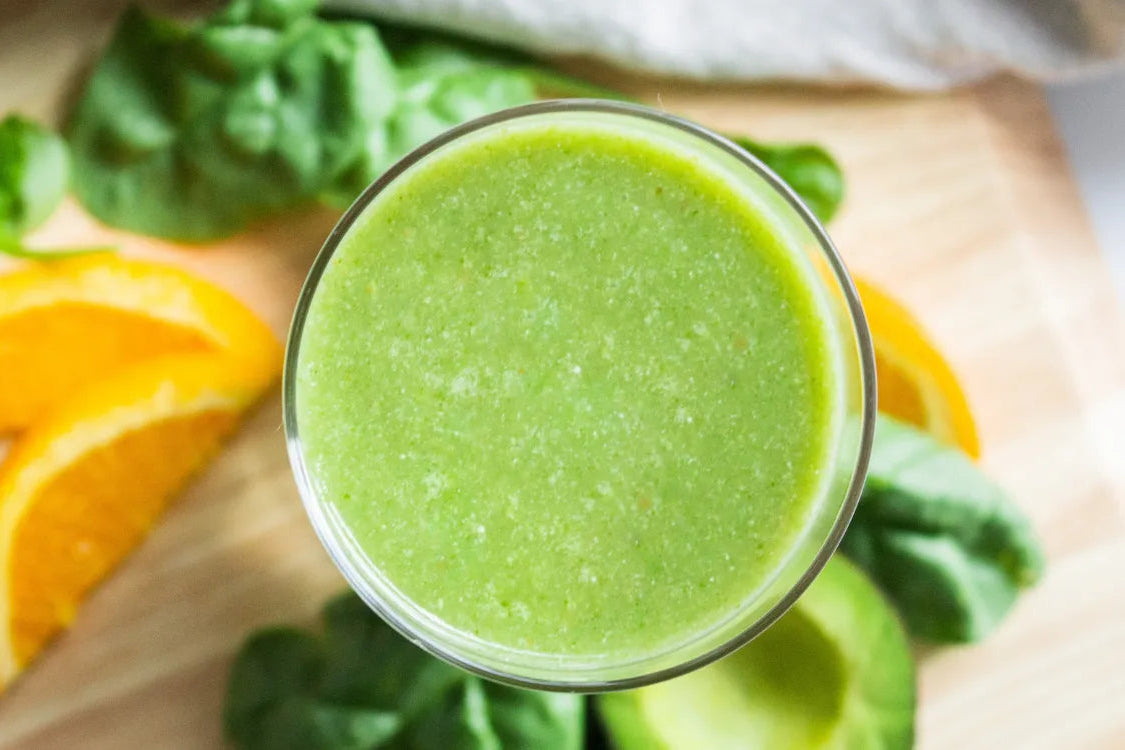 A healthy green smoothie surrounded by real ingredients like fresh spinach, oranges, and avocado.