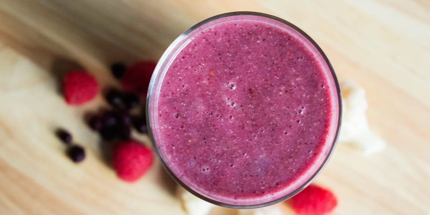 An overhead shot of a purple mixed-berry smoothie on a wooden table, surrounded by fresh fruit.