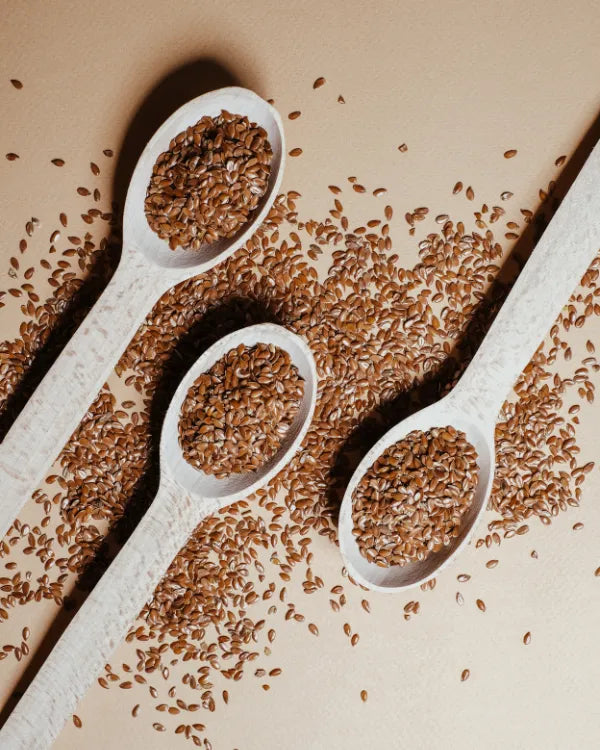 Flax seeds on three wooden spoons and scattered on a light brown surface.