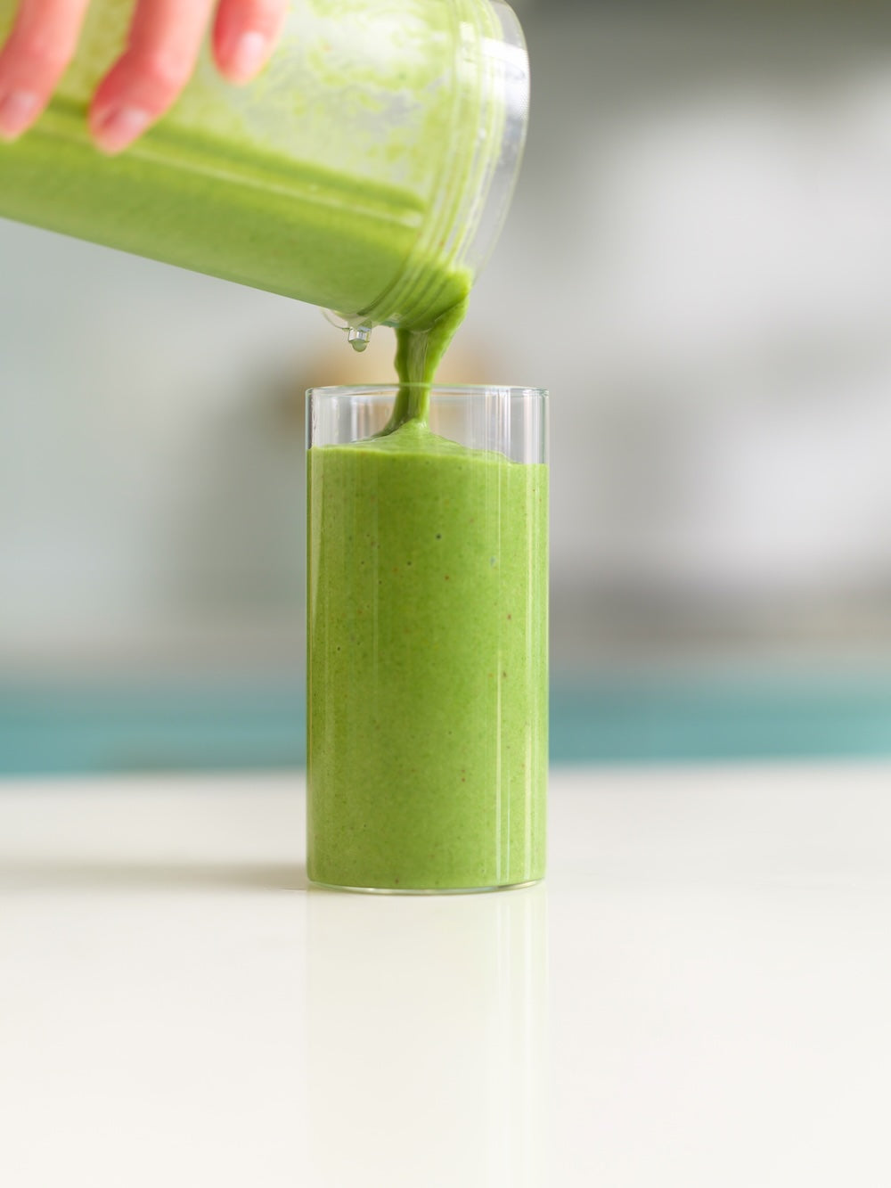 Green smoothie being poured from a blender into a glass on a white surface.