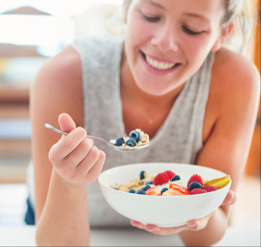 Smiling woman holding a spoon and a bowl of oats topped with fresh strawberries, raspberries, and blueberries