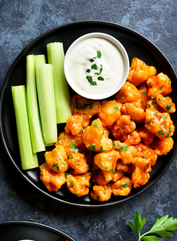Platter of buffalo cauliflower with celery and ranch dressing on a dark surface