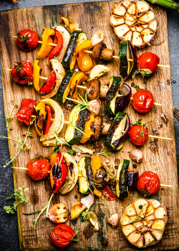 Grilled vegetable skewers on a wooden cutting board with grilled garlic bulbs