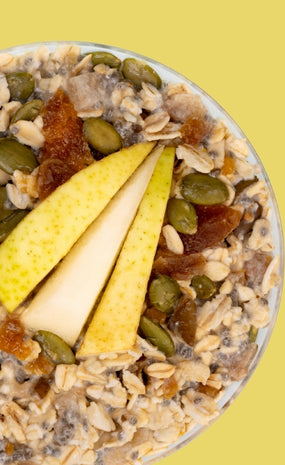 Overhead close-up of overnight oats topped with sliced pears, pumpkin seeds, and dried fruit pieces against a solid yellow background.