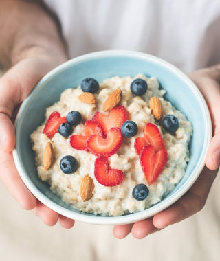 Close-up of hands holding a blue bowl of oats topped with blueberries, whole almonds, and strawberry slices cut into heart shapes.