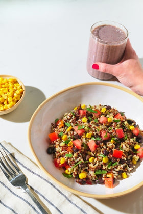 Colorful grain bowl with a hand holding a smoothie on a white surface