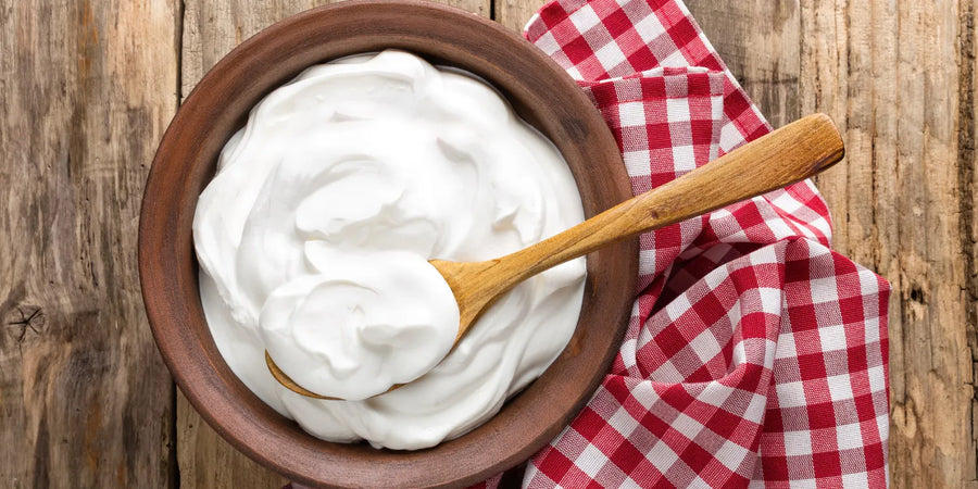 A top-down view of a rustic brown bowl filled with yogurt, with a wooden spoon resting inside, placed next to a red and white checkered cloth.